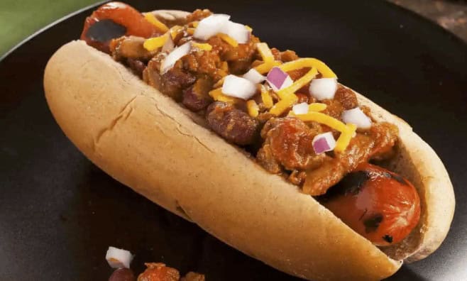 A grilled hot dog in a bun topped with chili, shredded cheese, and chopped onions, served on a dark plate—perfect for backyard barbecue flavor-packed recipes.